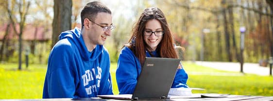 Two students sit at an outdoor table, deeply engaged with content on their laptop.